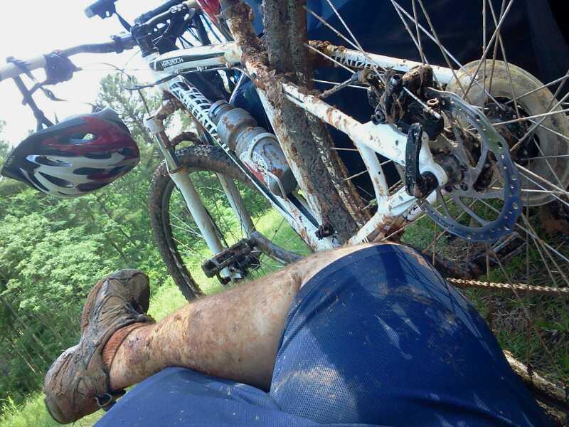 A close-up view of a muddy leg resting on the ground next to a mountain bike. The bike, partially obscured by a tree, is covered in dirt, indicating recent use on a muddy trail. In the background, trees and greenery suggest an outdoor setting. A bicycle helmet is visible on the bike. Buncombe - Brickhouse Recreation Area mountain bike trail.