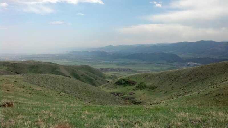 A panoramic view of a serene landscape featuring rolling green hills, with a backdrop of distant mountains under a partly cloudy sky. The foreground showcases lush grassy terrain, while the valley below stretches out towards the horizon, creating a peaceful and natural scene. Green Mountain mountain bike trail.