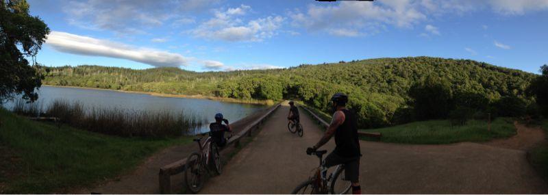 A panoramic view of a scenic landscape featuring three mountain bikers on a dirt path near a tranquil lake. Lush green hills surround the body of water, under a partly cloudy blue sky. The bikers, wearing helmets, are positioned on a bridge that stretches over the lake, with tall grasses and trees in the foreground. Annadel State Park mountain bike trail.