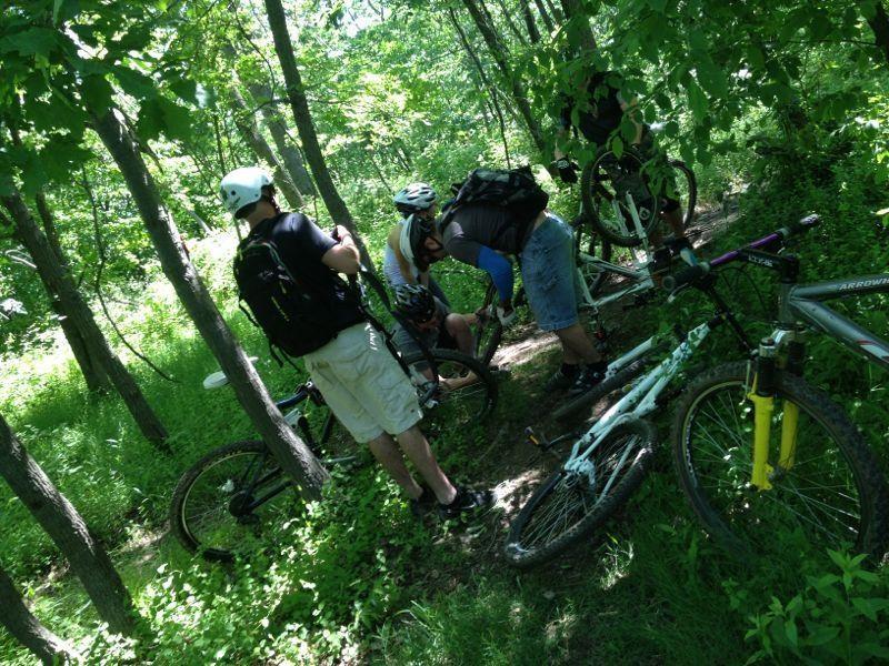 A group of four mountain bikers is gathered in a lush, green forest, attending to a bike on a narrow trail. Two bikes lie on the ground nearby, and the cyclists are wearing helmets and casual outdoor clothing. The scene captures the camaraderie of the group as they troubleshoot the bike issue amidst the vibrant foliage. Chimney Rock mountain bike trail.