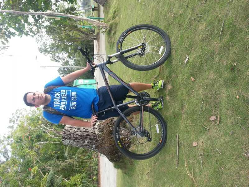 A person in a blue athletic shirt and black shorts stands beside a black mountain bike on a grassy area. The background features trees and a pathway, indicating an outdoor setting. The individual is smiling and holding the bike by its frame. Tortuguero mountain bike trail.