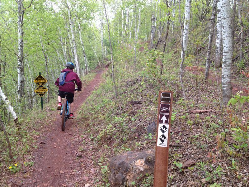A person riding a mountain bike on a dirt trail surrounded by green trees, with caution signs visible nearby indicating potential hazards ahead. The trail sign features symbols for biking and directions. Cuyuna Lakes mountain bike trail.