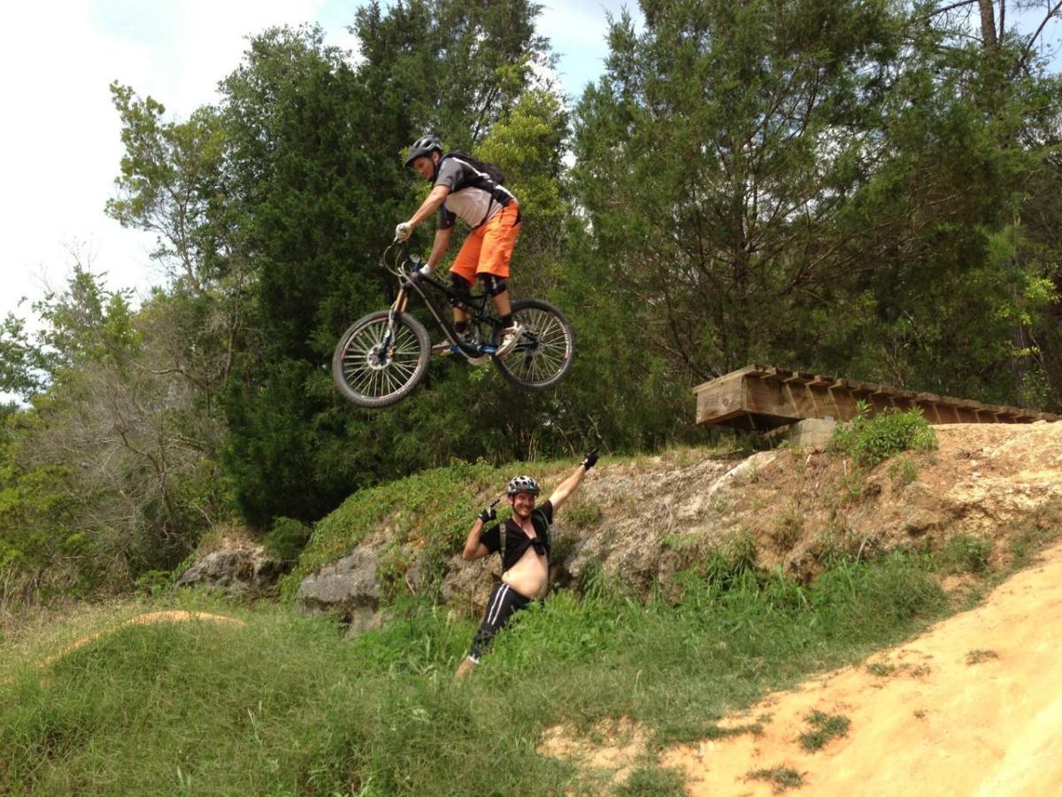 A mountain biker performing a jump over a dirt ramp, while another person watches from the ground, smiling and raising their hands in excitement. The scene is set in a green outdoor environment with trees in the background. Santos mountain bike trail.