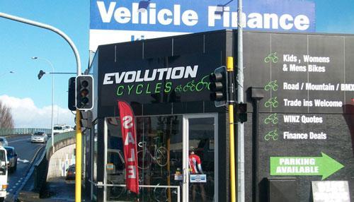 The image shows the exterior of a bike shop named "Evolution Cycles." The storefront features a large sign and a red "SALE" banner. Nearby, a traffic light is visible, and a vehicle finance sign is positioned above the shop. Additional information on the shop