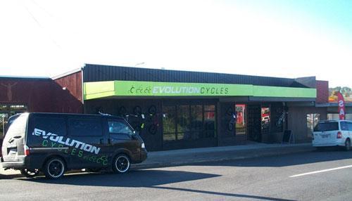 Exterior view of Evolution Cycles storefront, featuring a large sign with the shop's name in bright green letters. A dark-colored delivery van with the same branding is parked in front. The building is situated along a street, with additional vehicles visible in the background.