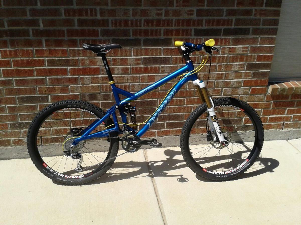 Turner 5.Spot: A blue mountain bike with yellow handlebars and a gold front suspension fork, parked on a concrete surface against a brick wall. The bike features thick tires and a rear derailleur, designed for off-road cycling.