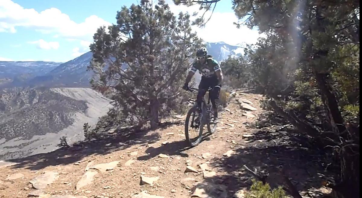 A mountain biker rides along a rocky trail surrounded by trees, with a scenic mountainous landscape in the background. The sky is blue with scattered clouds. Porcupine Rim mountain bike trail.