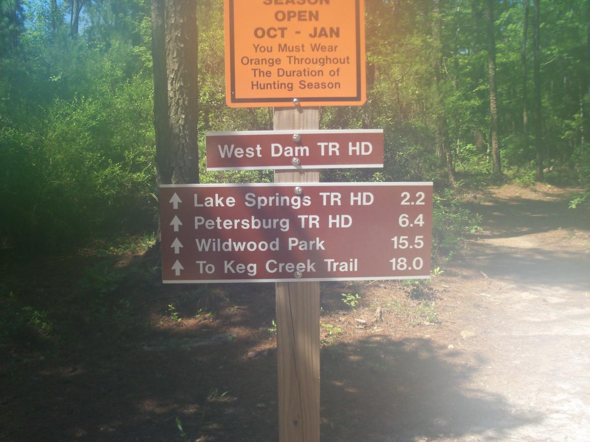A trail sign in a wooded area displaying directional arrows and distances to various locations: Lake Springs Trail Head (2.2 miles), Petersburg Trail Head (6.4 miles), Wildwood Park (15.5 miles), and Keg Creek Trail (18.0 miles). There is also a warning sign about the hunting season regulations. Bartram Trail / West Dam / Wildwood Park mountain bike trail.