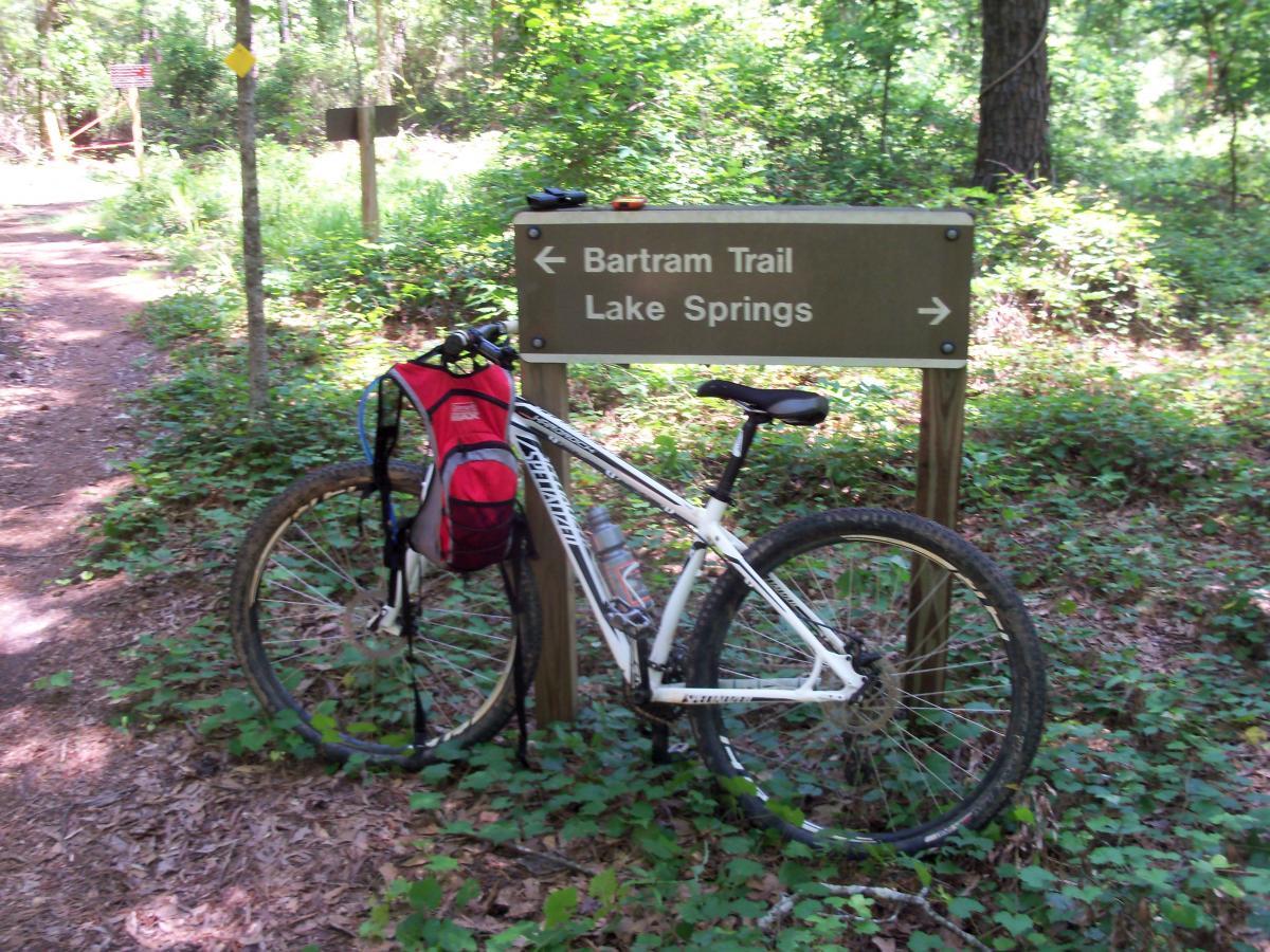 A mountain bike with a red backpack rests against a trail sign indicating directions to Bartram Trail and Lake Springs, surrounded by lush greenery and a dirt path in a wooded area. Bartram Trail / West Dam / Wildwood Park mountain bike trail.