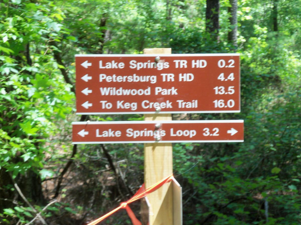 A wooden trail sign in a wooded area, displaying directions and distances to various hiking trails: Lake Springs TR HD (0.2 miles), Petersburg TR HD (4.4 miles), Wildwood Park (13.5 miles), To Keg Creek Trail (16.0 miles), and Lake Springs Loop (3.2 miles). The sign is surrounded by lush green foliage. Bartram Trail / West Dam / Wildwood Park mountain bike trail.
