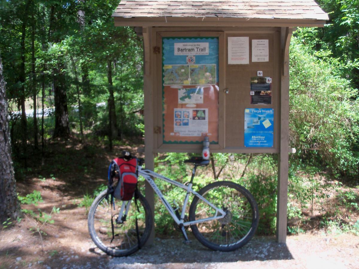 A mountain bike is parked beside a wooden information kiosk, which contains maps and trail guides for Bartram Trail. The kiosk is surrounded by lush green trees, and a water bottle is resting on the bike. The scene showcases a peaceful outdoor setting ideal for biking and hiking. Bartram Trail / West Dam / Wildwood Park mountain bike trail.