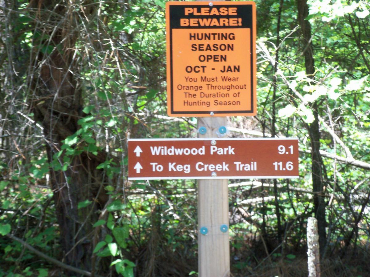 A wooden sign in a forested area displaying a warning about hunting season and directional information. The top part has an orange sign stating "PLEASE BEWARE! HUNTING SEASON OPEN OCT - JAN" with advice to wear orange clothing. Below, a brown sign points to "Wildwood Park" 9.1 miles away and "To Keg Creek Trail" 11.6 miles away. Green foliage surrounds the signs. Bartram Trail / West Dam / Wildwood Park mountain bike trail.