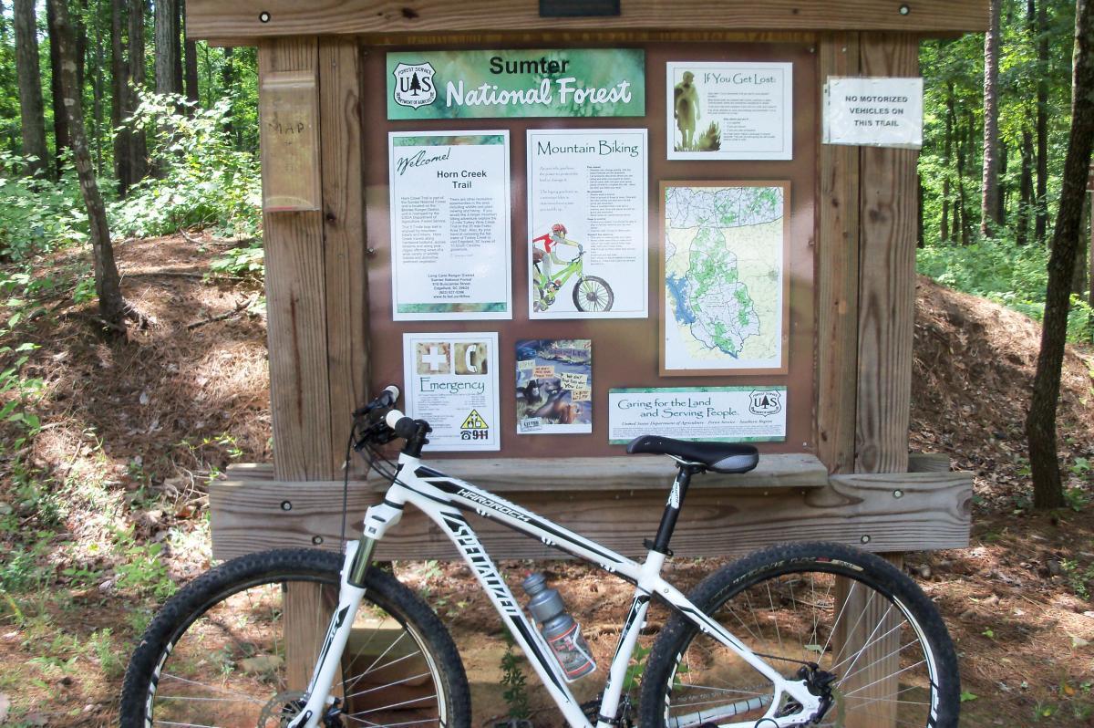 A wooden information kiosk in Sumter National Forest, featuring trail maps, guidelines for mountain biking, and emergency contact information. A mountain bike is parked in front of the kiosk, surrounded by trees and greenery. The sign emphasizes the Horn Creek Trail and includes important notices, such as restrictions on motorized vehicles. Lick Fork (Horn Creek) mountain bike trail.