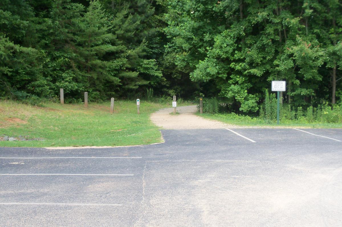 Parking lot at the edge of a forested area, with a gravel path leading into the trees. There are wooden posts along the path, and signage indicating regulations or information about the area. The surroundings are green and lush, suggesting a natural setting for outdoor activities. Colonel Francis Beatty Park mountain bike trail.