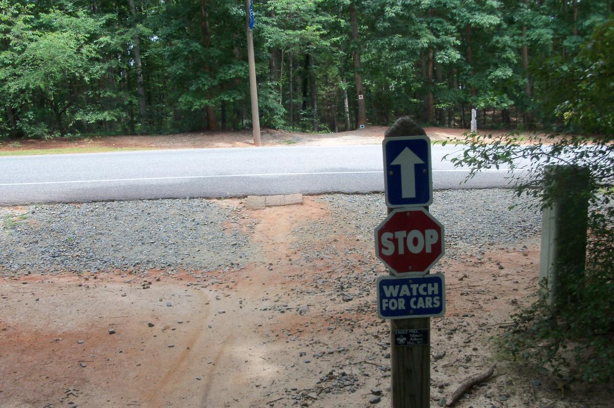 A stop sign and a sign that says "Watch for Cars" are positioned near a gravel entrance to a road. In the background, a paved road runs parallel, surrounded by dense trees. The scene is set in a natural outdoor environment. Colonel Francis Beatty Park mountain bike trail.