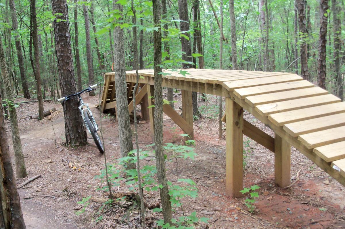 A trail in a wooded area featuring a wooden bike ramp curved over the ground, with a mountain bike leaned against a tree nearby. Lush green foliage surrounds the path, and the ground is covered in brown dirt and scattered leaves. Colonel Francis Beatty Park mountain bike trail.