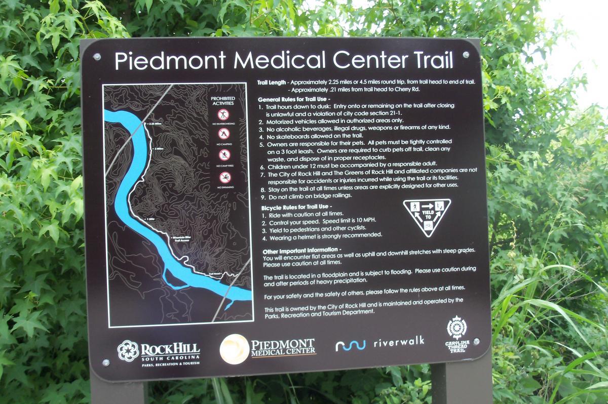 Signage for the Piedmont Medical Center Trail detailing trail length, rules for use, bicycle guidelines, and safety information, set against a backdrop of greenery. River walk mountain bike trail.