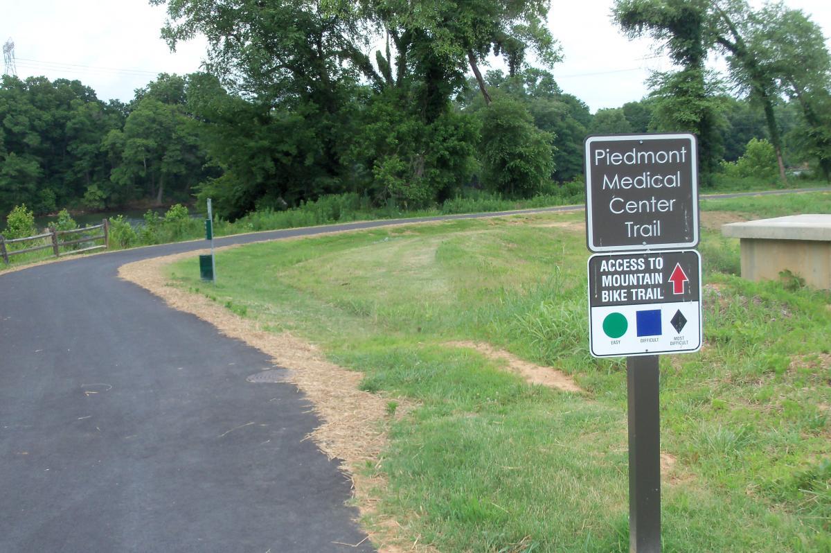 Sign indicating the Piedmont Medical Center Trail and access to a mountain bike trail, with a paved pathway lined by grass and trees in the background. River walk mountain bike trail.