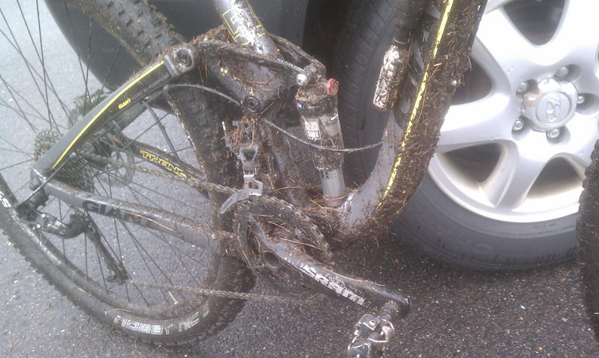 Giant Trance X 29er: A close-up view of a muddy mountain bike, showcasing the dirty frame and drivetrain, with mud and debris covering the components. In the background, part of a car tire is visible on a gravel surface.