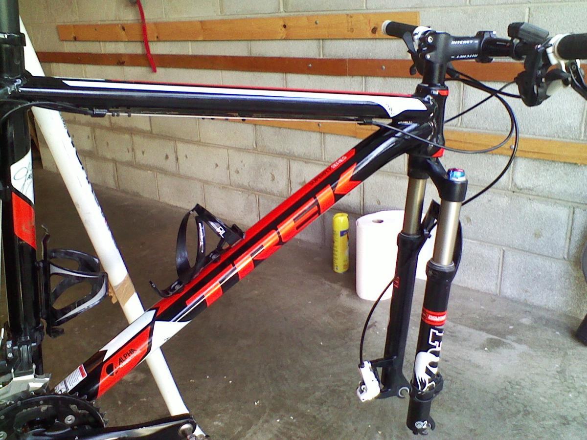 Trek Superfly 100 AL: A close-up view of a black and red Trek mountain bike frame, featuring detailed components such as the handlebars, suspension fork, and water bottle cage. The bike is set against a backdrop of a garage with concrete walls and wooden shelves.