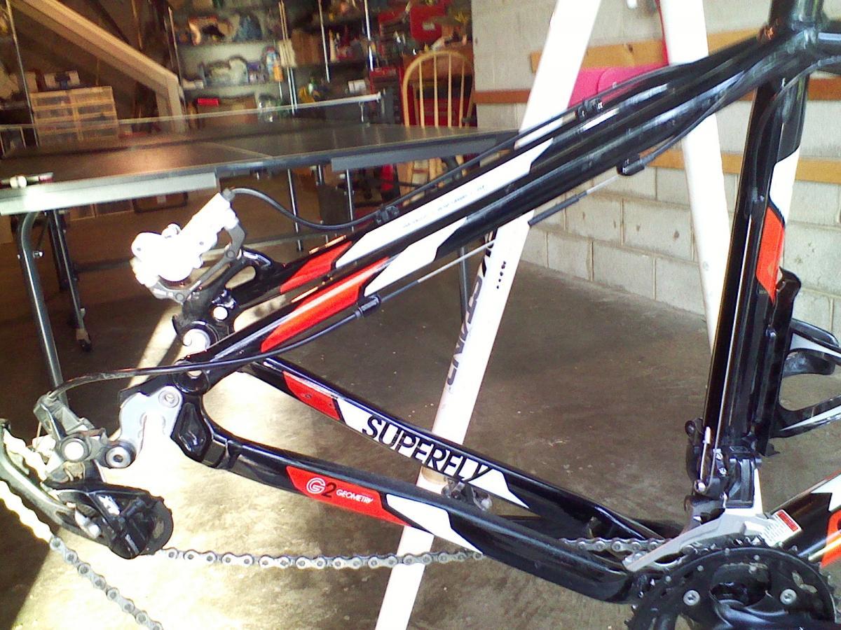 A close-up view of a bicycle frame, specifically a "Superfly" model, displayed in a garage setting. The image captures the bike's drivetrain and rear suspension components, with a chain and derailleur visible. In the background, a ping pong table and shelves are partially seen, indicating a domestic environment.