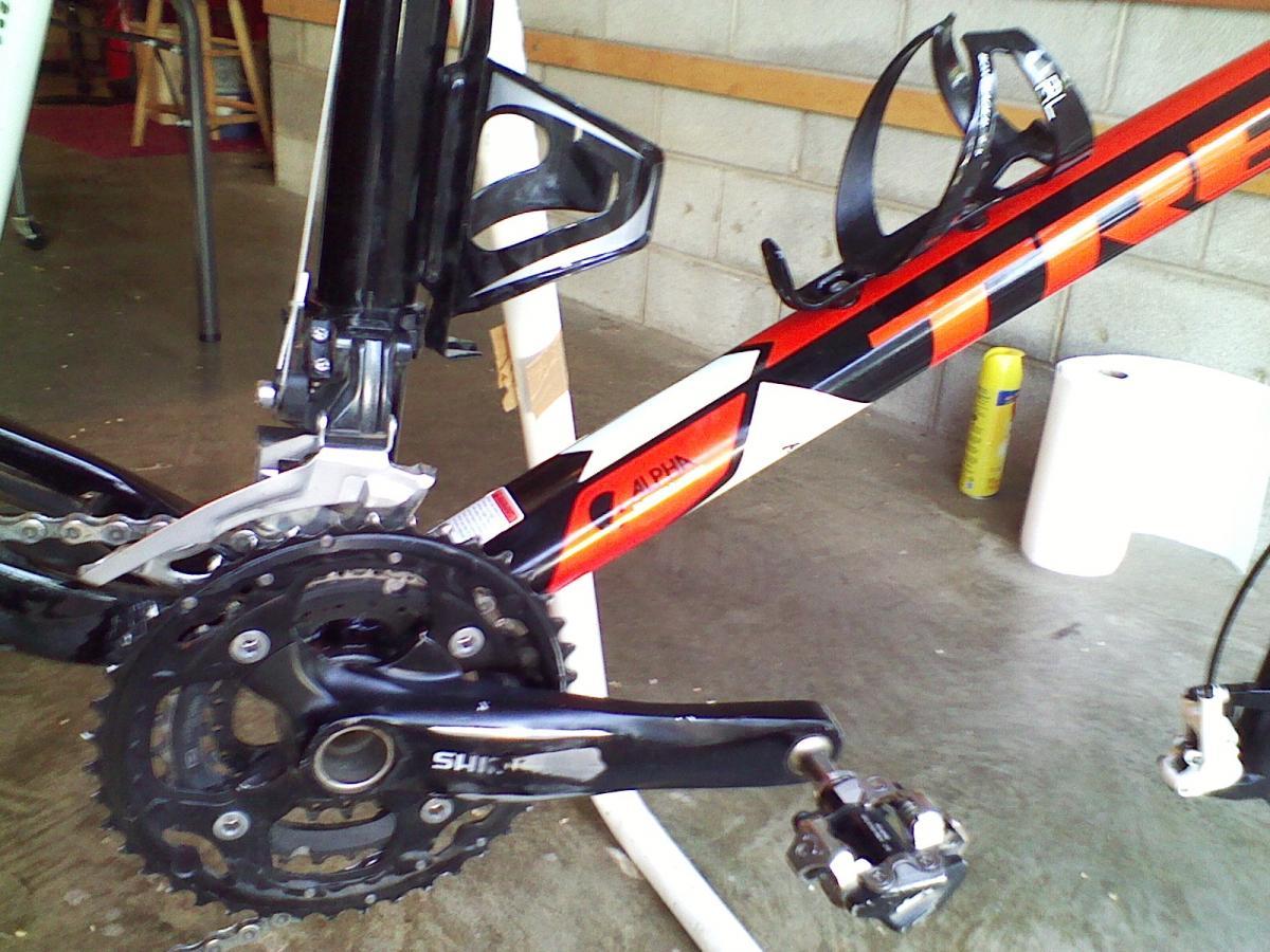 Trek Superfly 100 AL: Close-up view of the drivetrain and bottom bracket area of a mountain bike, featuring a black and red frame, crankset, chain, and pedal. The background shows a garage environment with concrete flooring and a few items in the background.