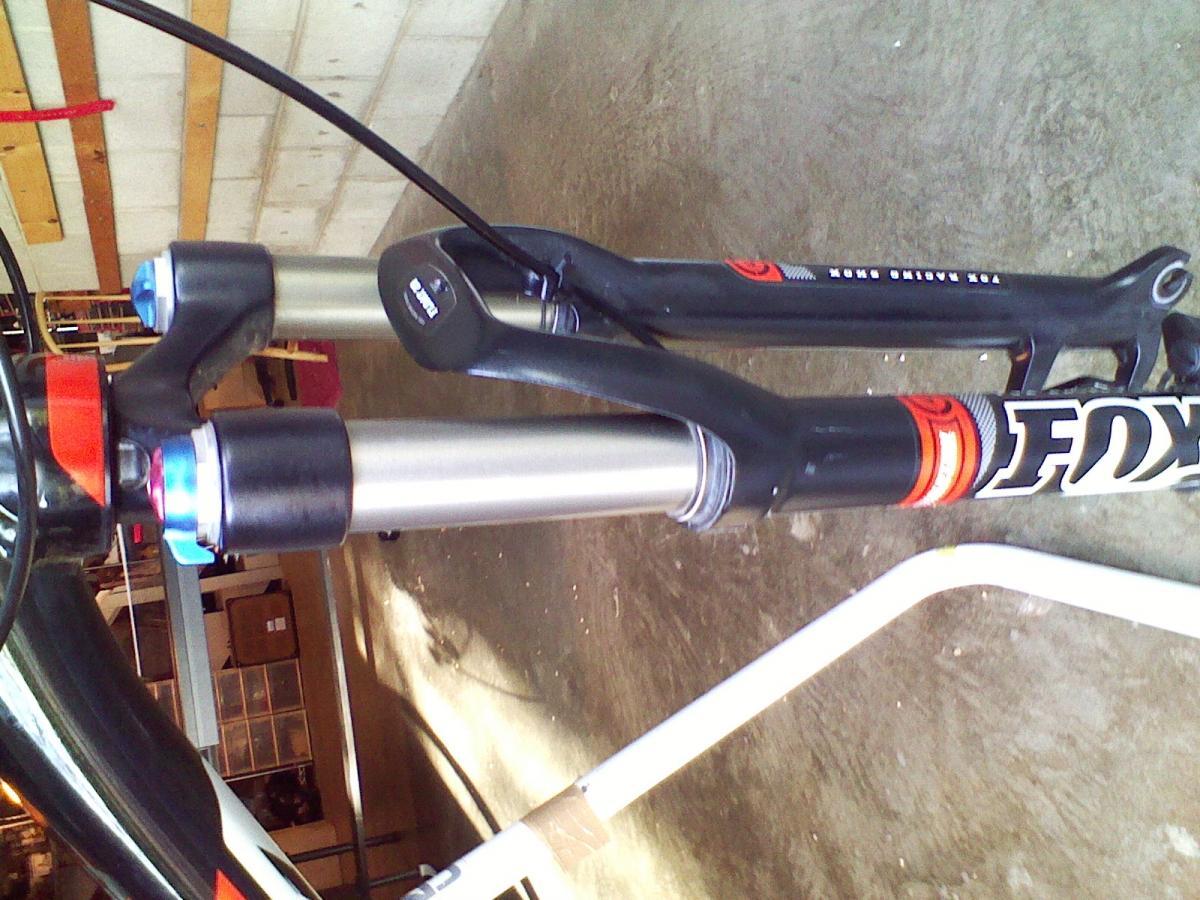 Trek Superfly 100 AL: Close-up view of a mountain bike front fork, specifically showing the suspension components. The fork features labeled adjustment knobs in blue and red, along with the branding "FOX" on the side. The background includes a garage setting with tools and storage visible, highlighting a workspace for bike maintenance.