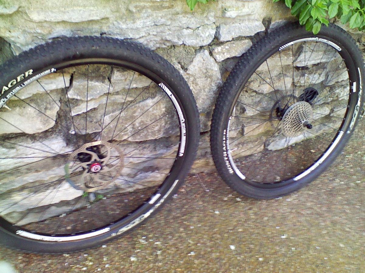 Trek Superfly 100 AL: Two mountain bike wheels resting against a stone wall. The wheels are positioned side by side, showing off their thick tires and disc brakes. The background features a mix of stone and greenery.