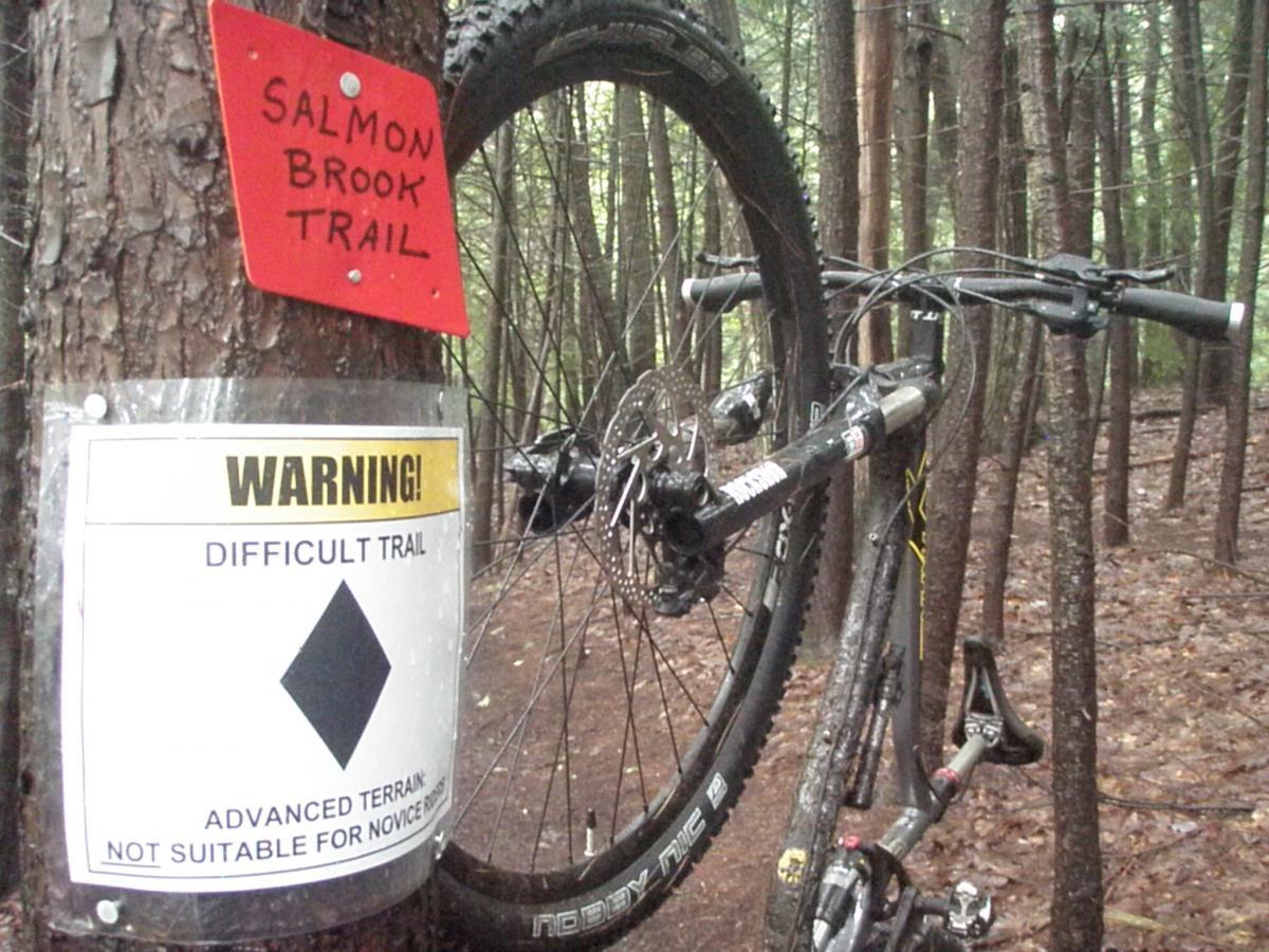 A mountain bike leaning against a tree on a forest trail, with a red sign reading "Salmon Brook Trail" above and a warning sign indicating a "Difficult Trail" with a diamond symbol. The area is surrounded by dense trees and damp ground, suggesting a rustic and challenging terrain for biking. Franklin Falls mountain bike trail.