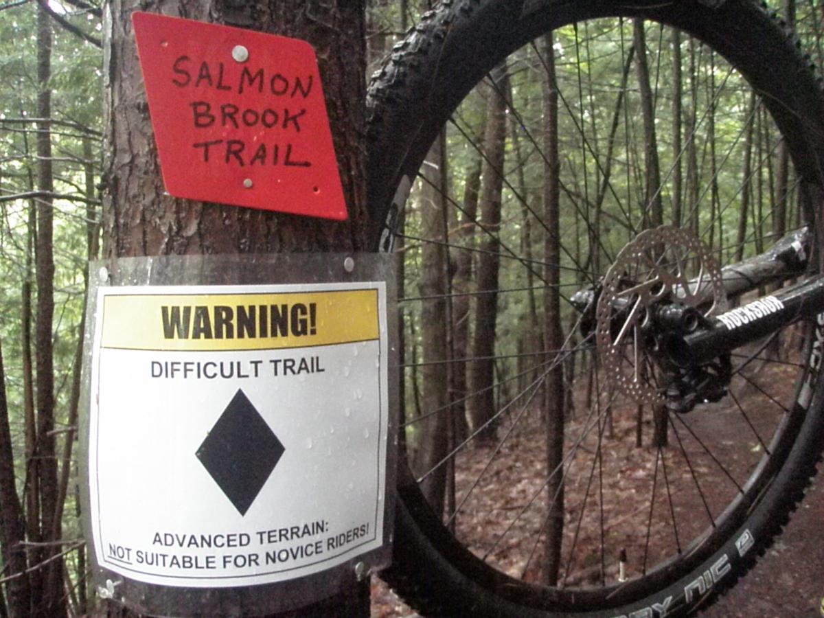A close-up view of a mountain bike wheel leaning against a tree on a forest trail. Above the wheel, there is a red sign labeled "Salmon Brook Trail." Below, a warning sign states "WARNING! DIFFICULT TRAIL" with a diamond symbol, indicating that the trail features advanced terrain not suitable for novice riders. The background consists of dense green foliage, suggesting a wooded environment. Franklin Falls mountain bike trail.