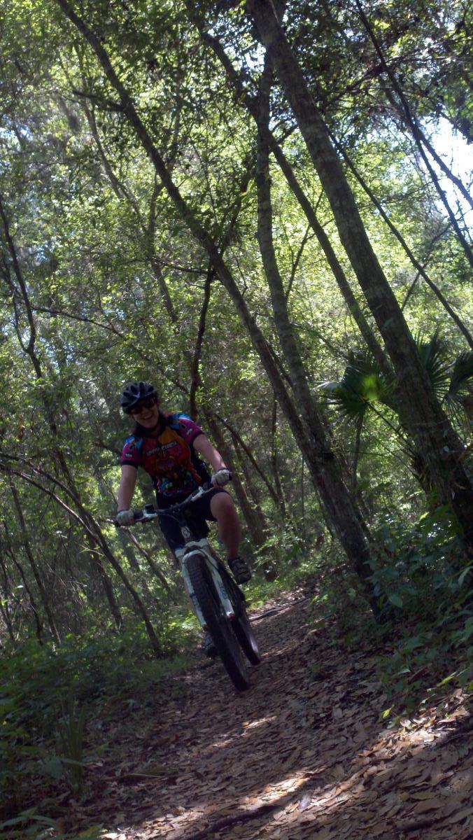 A person riding a mountain bike on a trail through a lush green forest, wearing a colorful cycling jersey and a helmet, smiling as they navigate the pathway surrounded by trees and foliage. Santos mountain bike trail.