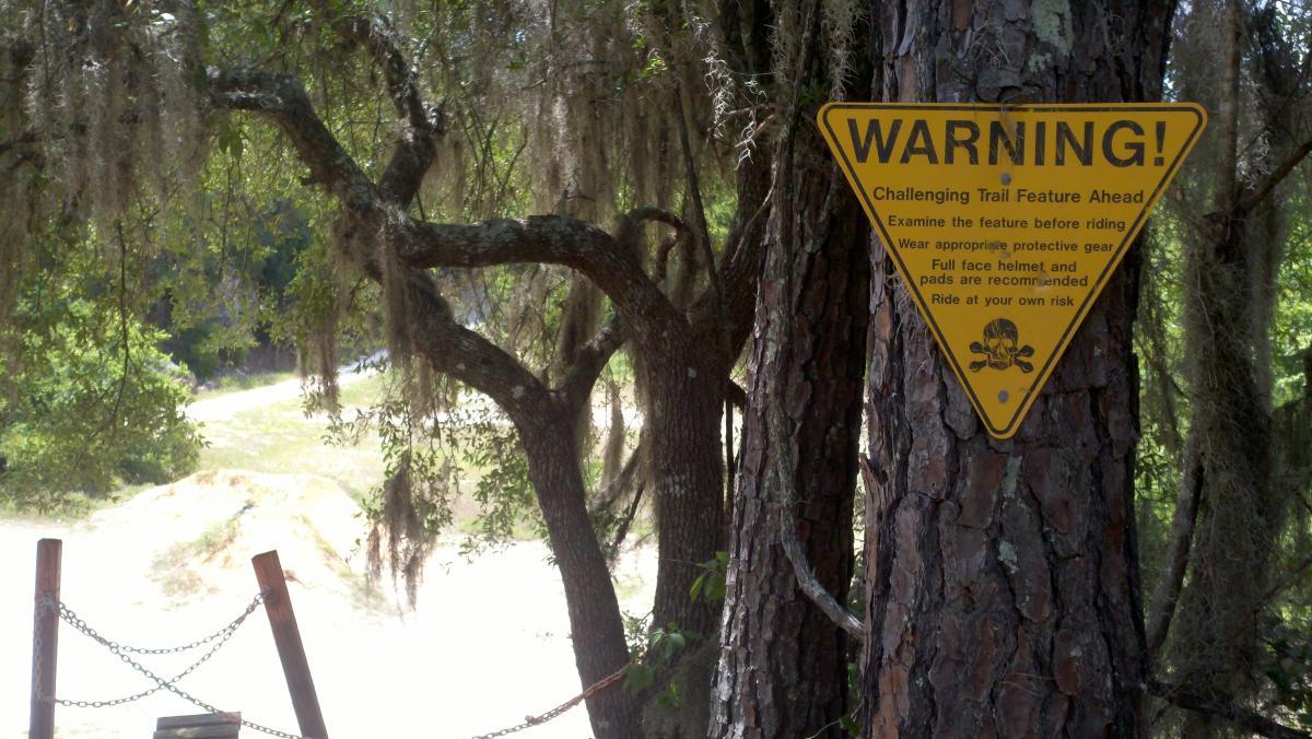 A warning sign affixed to a tree, indicating a challenging trail feature ahead. The sign advises riders to examine the feature before riding, wear appropriate protective gear, including a full-face helmet and pads, and to ride at their own risk. Green foliage and a dirt path can be seen in the background. Santos mountain bike trail.