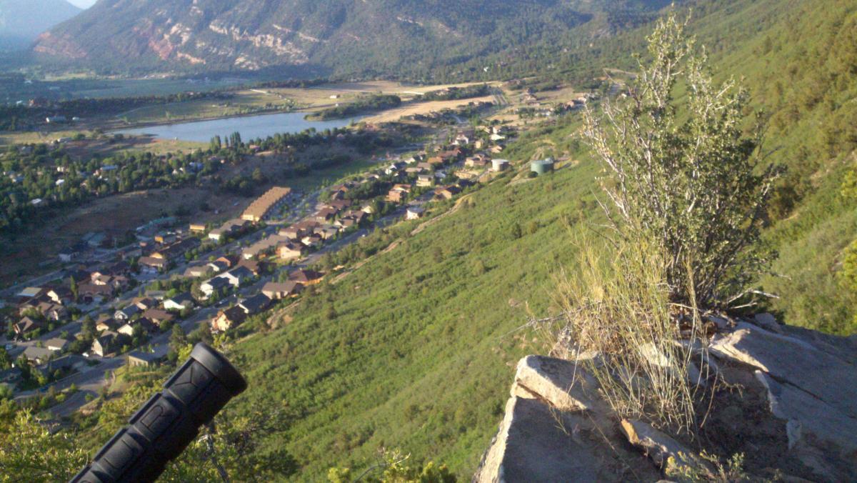 A panoramic view of a valley surrounded by mountains, featuring a small lake and residential houses nestled on the hillsides. In the foreground, a rocky outcrop with a shrub stands prominently, while the landscape stretches out below with greenery and homes visible. Horse Gulch mountain bike trail.