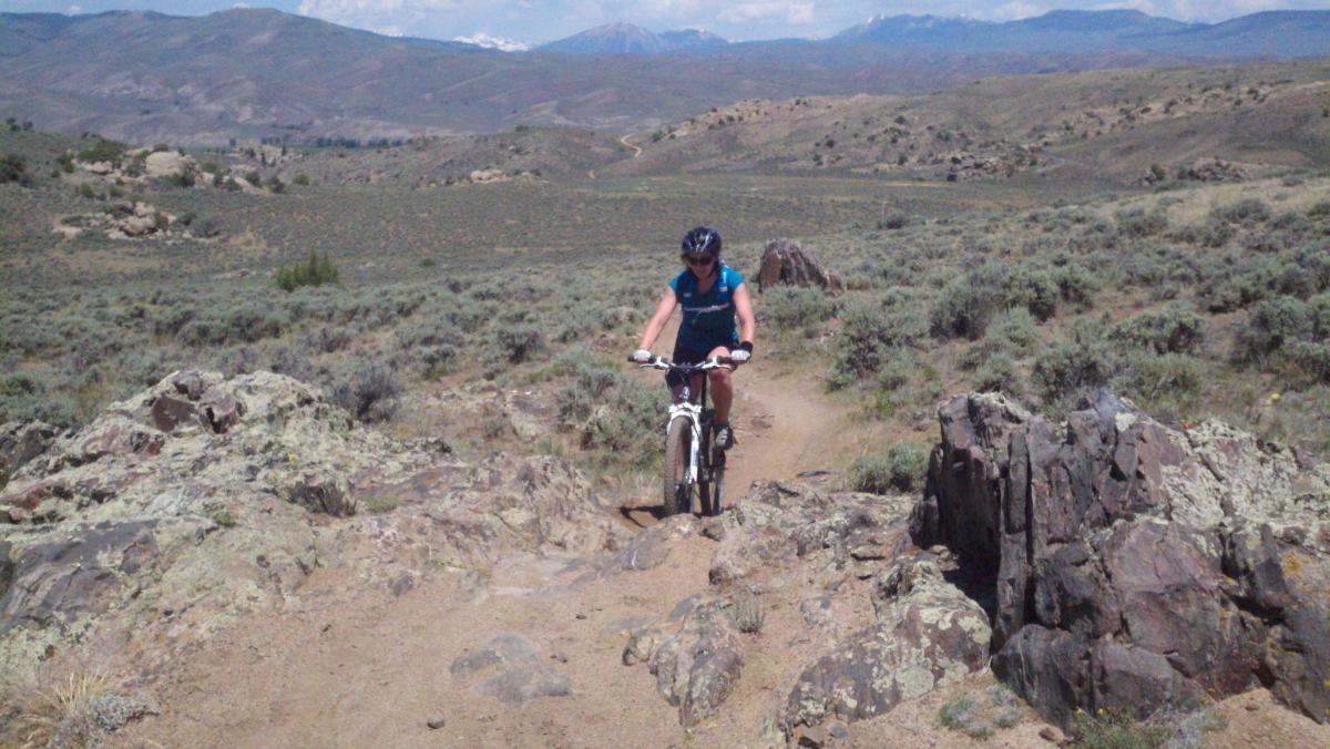 A cyclist navigating a rocky trail in a mountainous landscape, surrounded by shrubs and rolling hills under a clear blue sky. Hartman Rocks mountain bike trail.