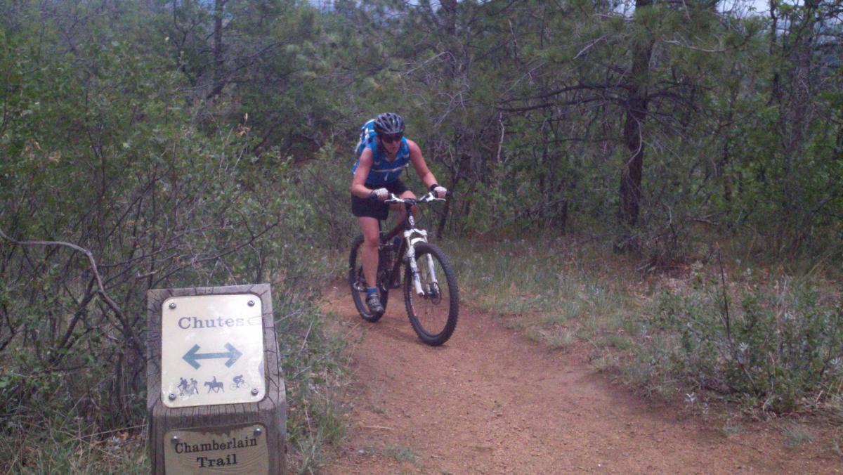 A mountain biker navigating a dirt trail surrounded by lush greenery, with a trail sign indicating directions to "Chutes" and "Chamberlain Trail." The biker is wearing a blue jersey and helmet, focused on the path ahead. Stratton Open Space / The Chutes mountain bike trail.