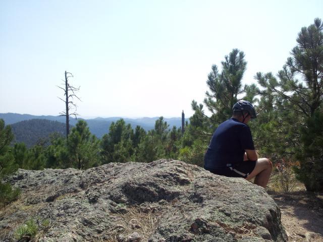 A person wearing a helmet sits on a rock in a forested mountainous area, overlooking a scenic view of hills and trees under a clear blue sky. Centennial Trail mountain bike trail.