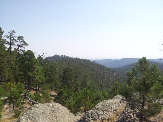 A panoramic view of a mountainous landscape featuring dense forested hills, scattered rocks in the foreground, and a hazy sky. The scene captures the natural beauty and tranquility of the wilderness. Centennial Trail mountain bike trail.