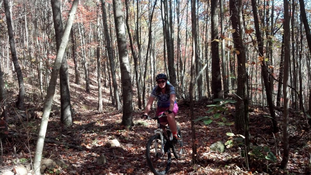 A person riding a mountain bike along a forest trail, surrounded by trees with autumn leaves. The cyclist wears a helmet and smiles as they navigate the path. Sunlight filters through the trees, creating a vibrant, natural atmosphere. Tanasi Trail System mountain bike trail.
