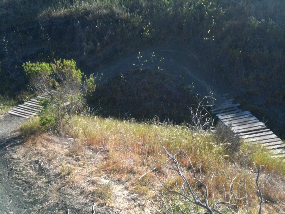 A view of a natural landscape featuring a grassy area and two small wooden bridges spanning a narrow ravine or ditch. The scene includes sparse vegetation and dry grass, with sunlight illuminating the surroundings. San Clemente Singletracks mountain bike trail.