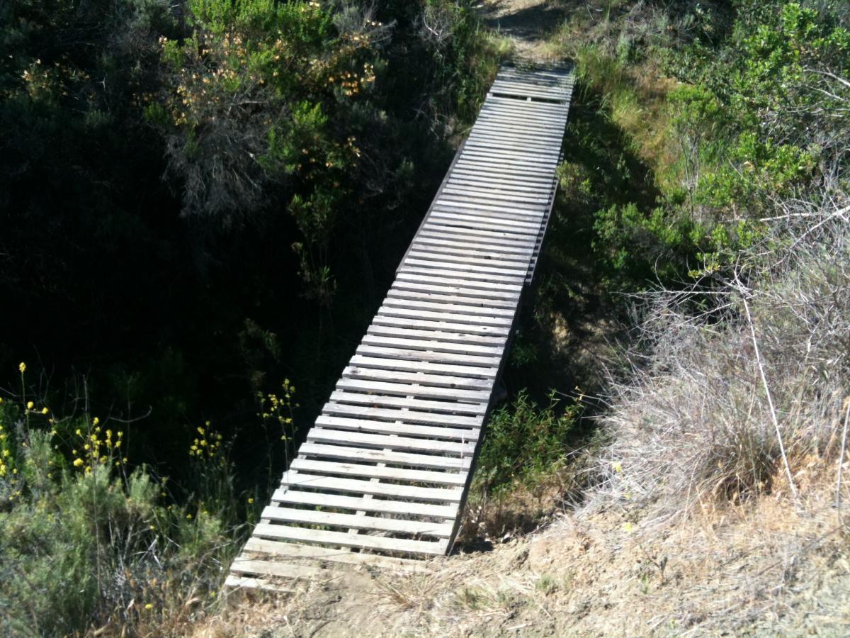 Alt text: A wooden bridge made of slatted planks spans a small ravine, surrounded by green shrubs and dry grass. The bridge leads from one side of the landscape to the other, connecting two elevated areas. San Clemente Singletracks mountain bike trail.