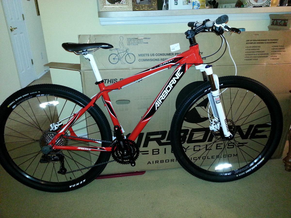 Airborne Guardian: A red Airborne mountain bike is displayed in a room, next to its packaging box. The bike features large black tires with white lettering, a black seat, and a white front fork. The background shows a doorway and various home decor elements.