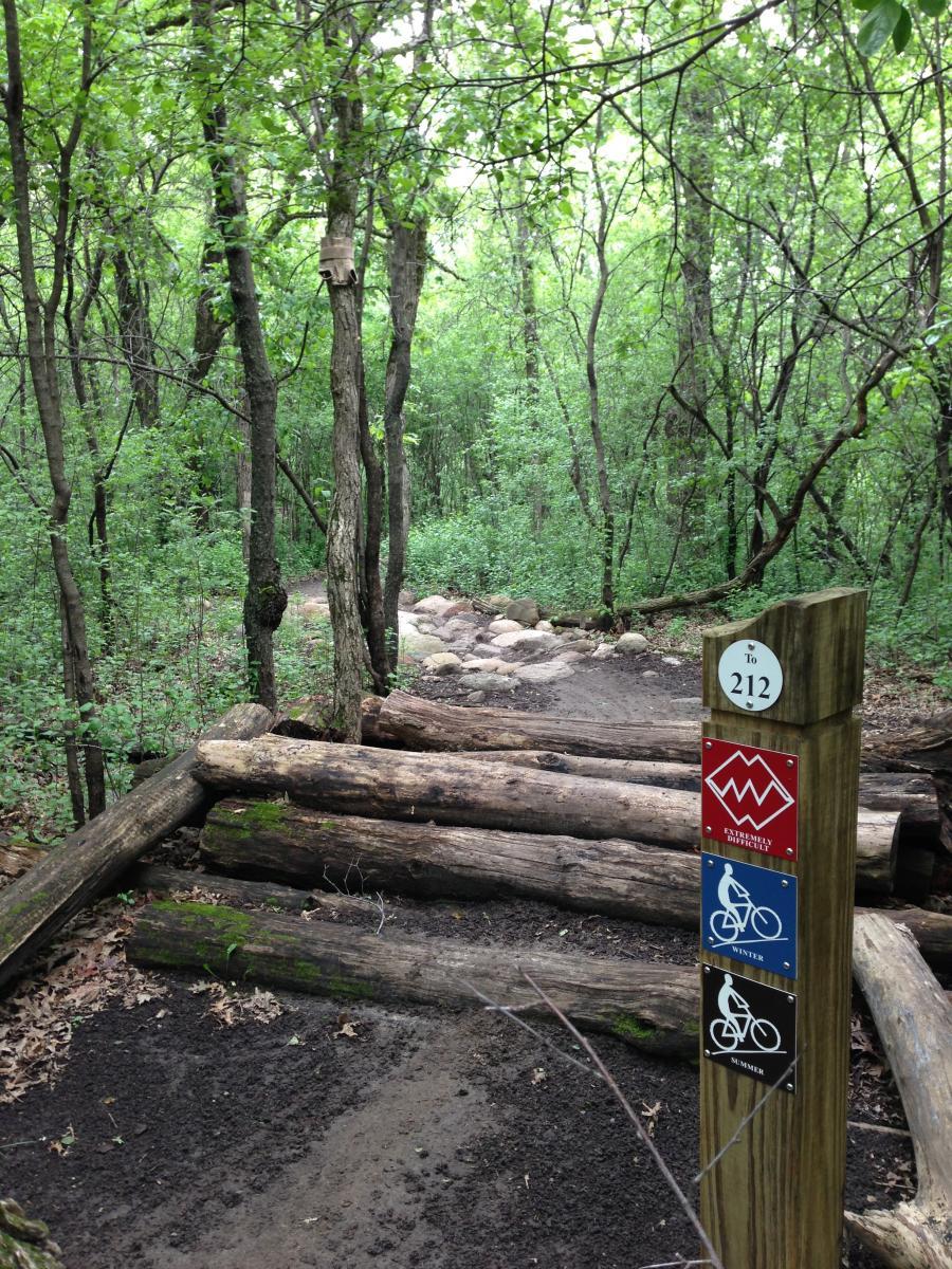 A wooded trail entrance with a signpost indicating trail difficulty and usage for biking, featuring three colored signs for "Extremely Difficult," "Winter," and "Summer." The path is marked by logs and stones, surrounded by lush green trees and foliage. Lebanon Hills mountain bike trail.