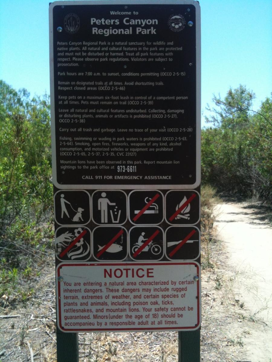 A sign at Peters Canyon Regional Park providing park regulations and safety information. The sign includes park hours, guidelines for wildlife and plant preservation, safety warnings, and prohibited activities, accompanied by icons illustrating these rules. Plant life and natural surroundings can be seen in the background. Peters Canyon Regional Park mountain bike trail.