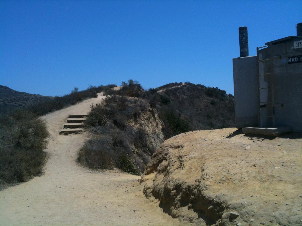 A dirt path leads uphill with stone steps on the left side, surrounded by sparse vegetation and hills under a clear blue sky. To the right, there is a concrete structure with industrial features, situated on the edge of the hilltop. Peters Canyon Regional Park mountain bike trail.