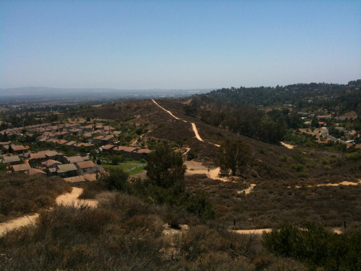A panoramic view from a hillside overlooking a residential area with houses and green spaces below, surrounded by gentle rolling hills and distant mountains under a clear blue sky. A dirt path winds through the landscape, leading into the distance. Peters Canyon Regional Park mountain bike trail.