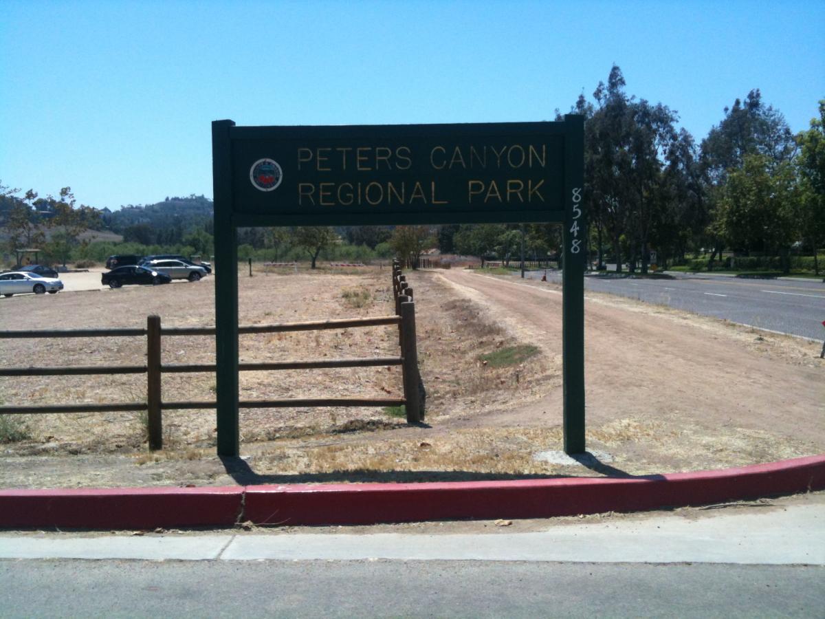 Sign for Peters Canyon Regional Park, displaying the park's name prominently, with a dirt area and parking spaces visible nearby. The background features trees and a clear blue sky. Peters Canyon Regional Park mountain bike trail.