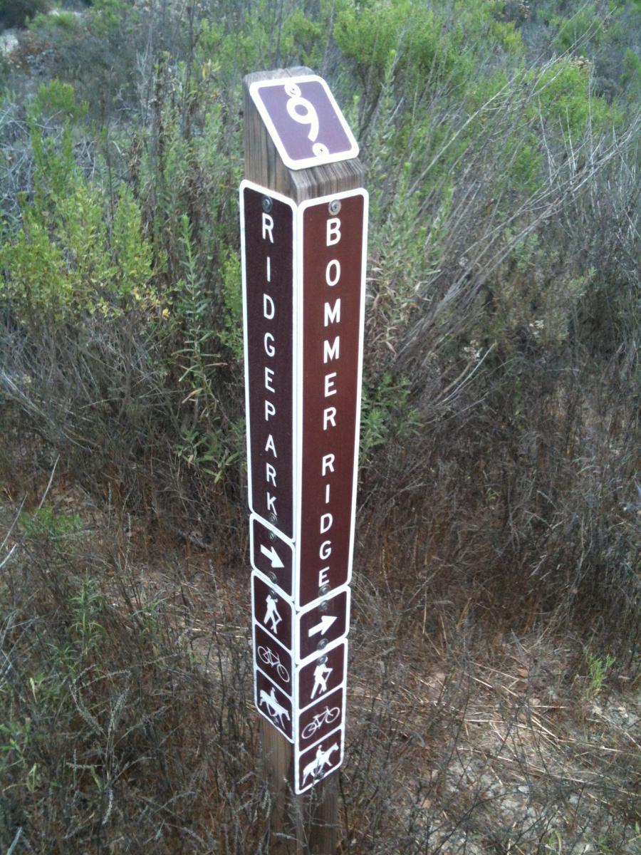 Signpost at a trail intersection indicating directions for Ridge Park and Bommer Ridge, featuring icons for hiking, biking, and dog walking, with the number 9 displayed at the top. Surrounding foliage consists of shrubs and plants typical of a natural landscape. Gate 9 mountain bike trail.