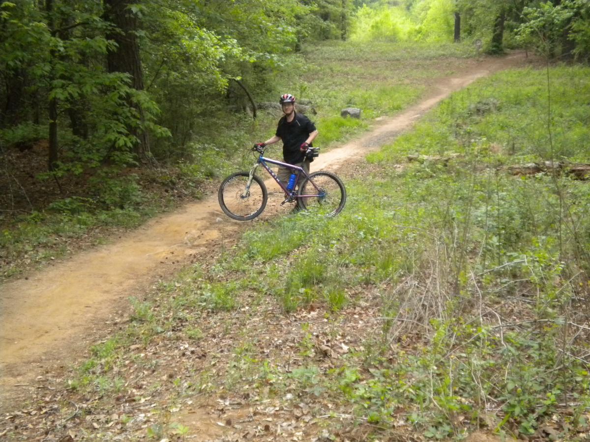 A person wearing a helmet stands next to a mountain bike on a dirt trail surrounded by lush greenery and trees. The path diverges into two directions, indicating a biking route through a forested area. Fort Yargo State Park mountain bike trail.