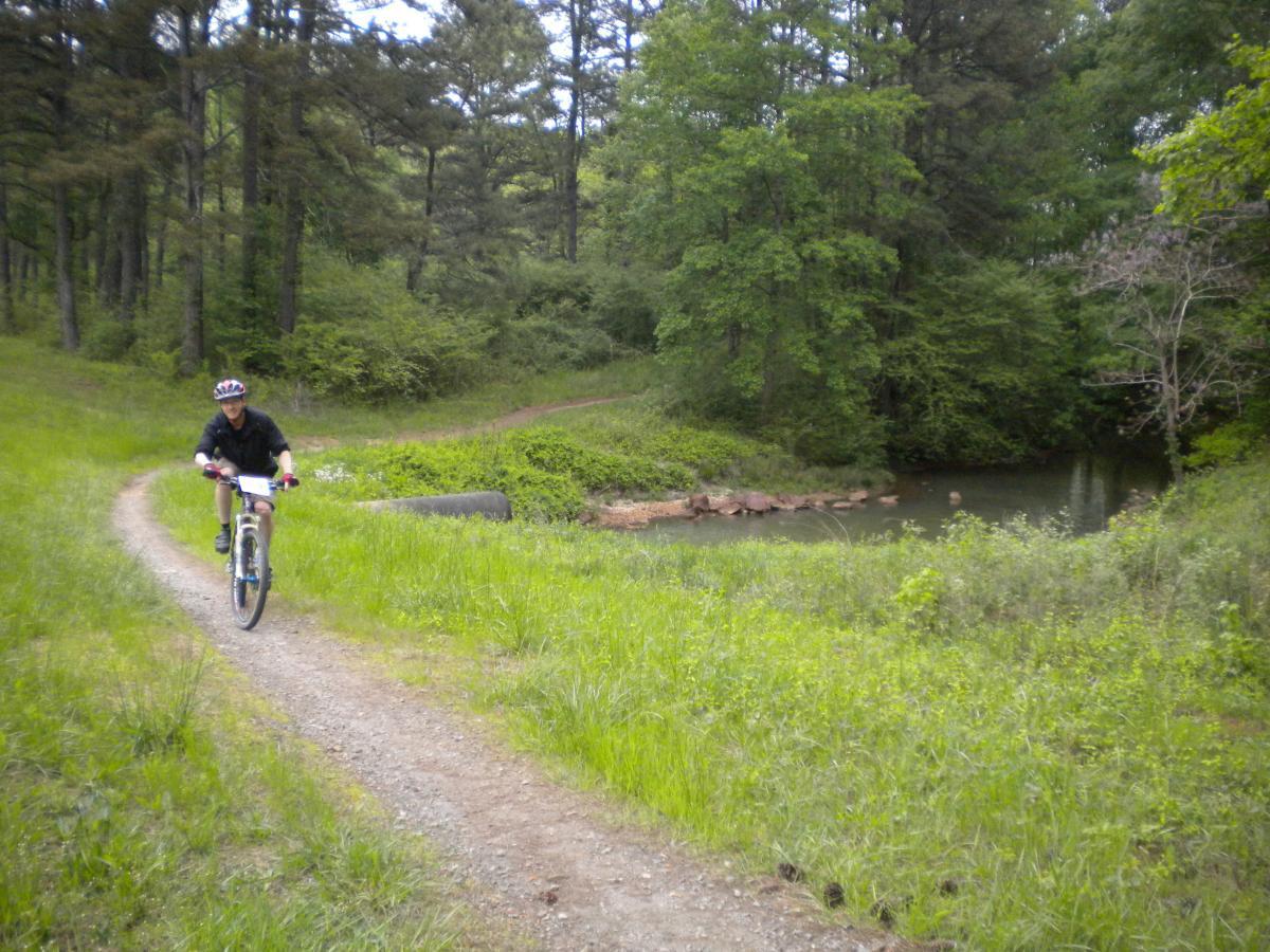 A person riding a mountain bike on a gravel path through a lush, green forest. The background features trees and a small pond, with vibrant foliage surrounding the trail. Fort Yargo State Park mountain bike trail.
