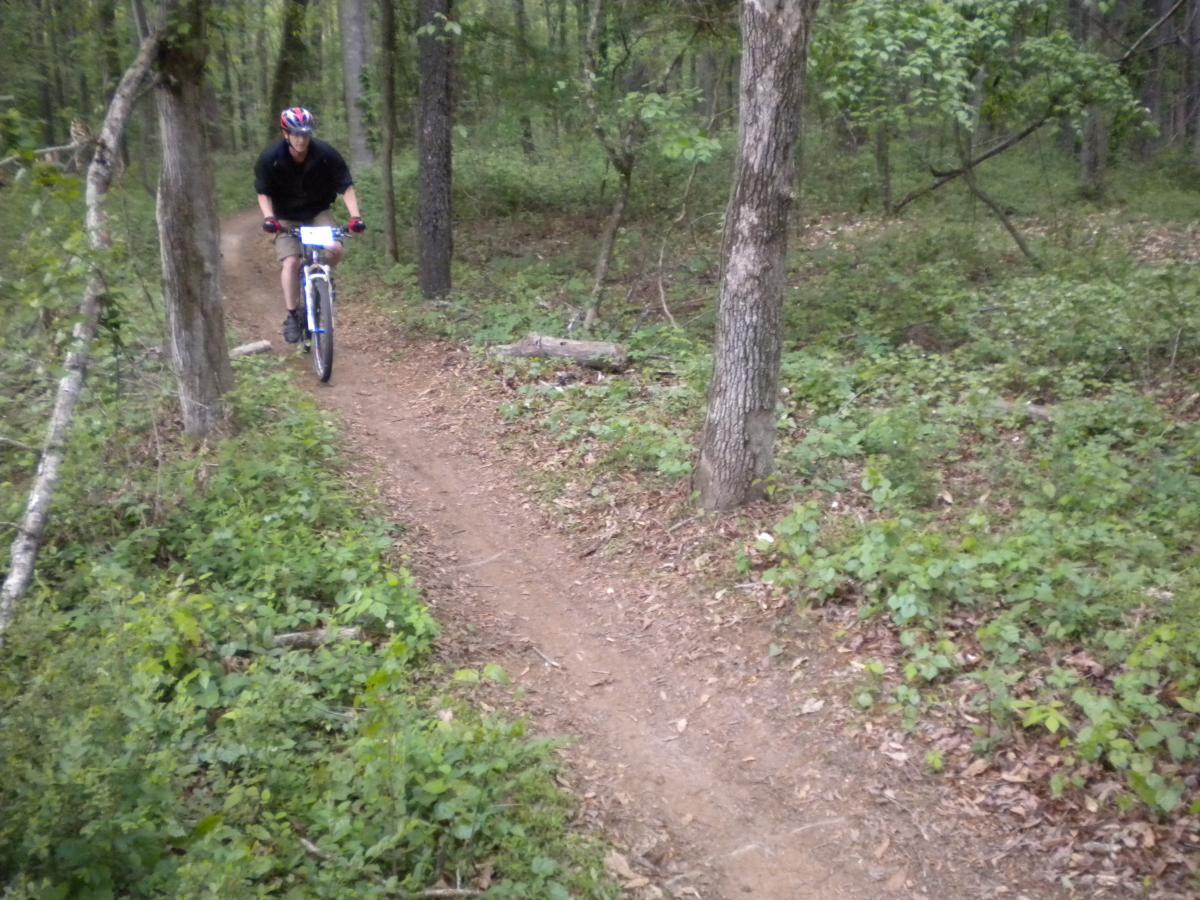 A cyclist riding along a narrow dirt trail in a wooded area, surrounded by trees and greenery. The cyclist is wearing a helmet and riding a mountain bike. Fort Yargo State Park mountain bike trail.